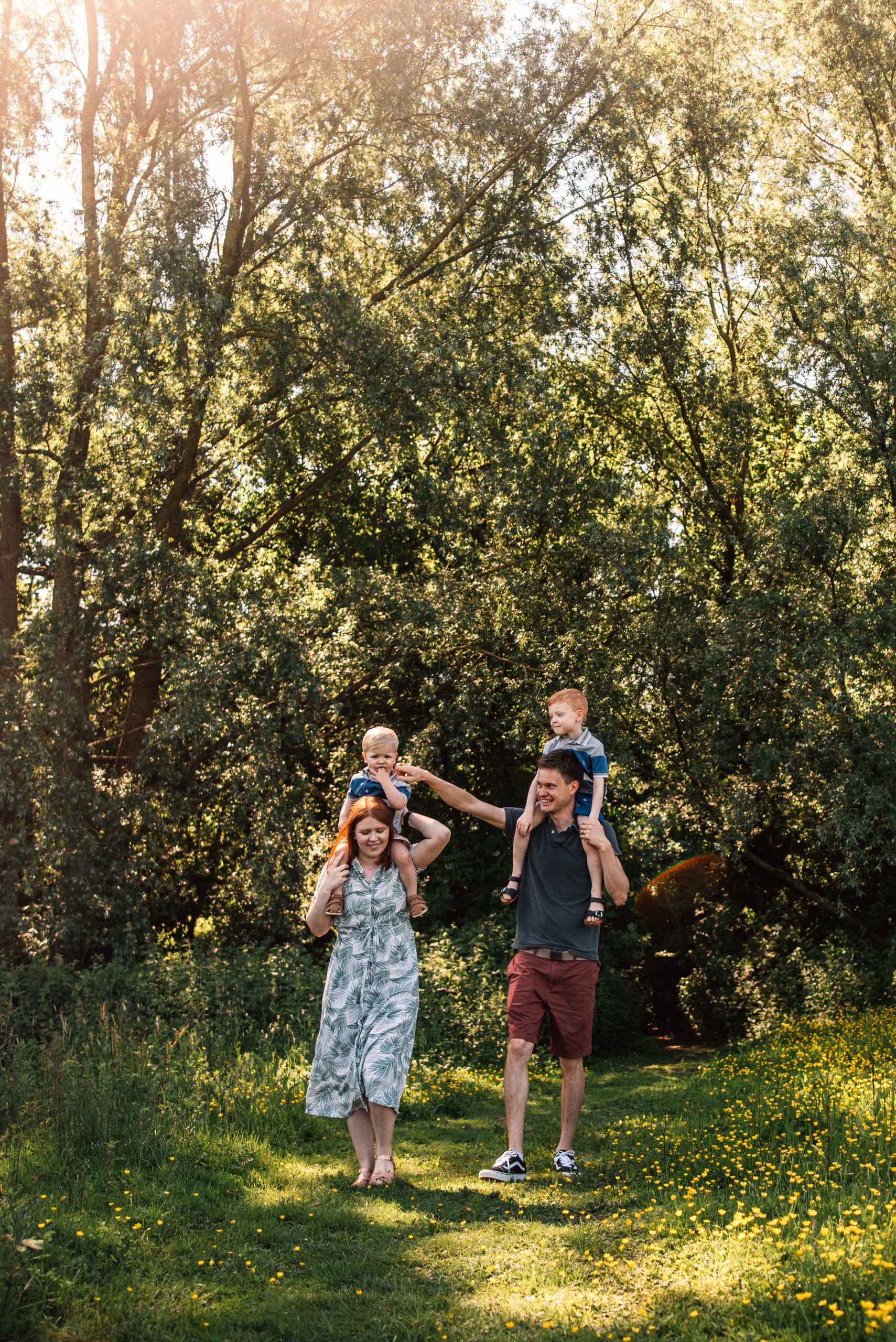 mum and dad each hold a child on their shoulders as they walk in a sunlit meadow