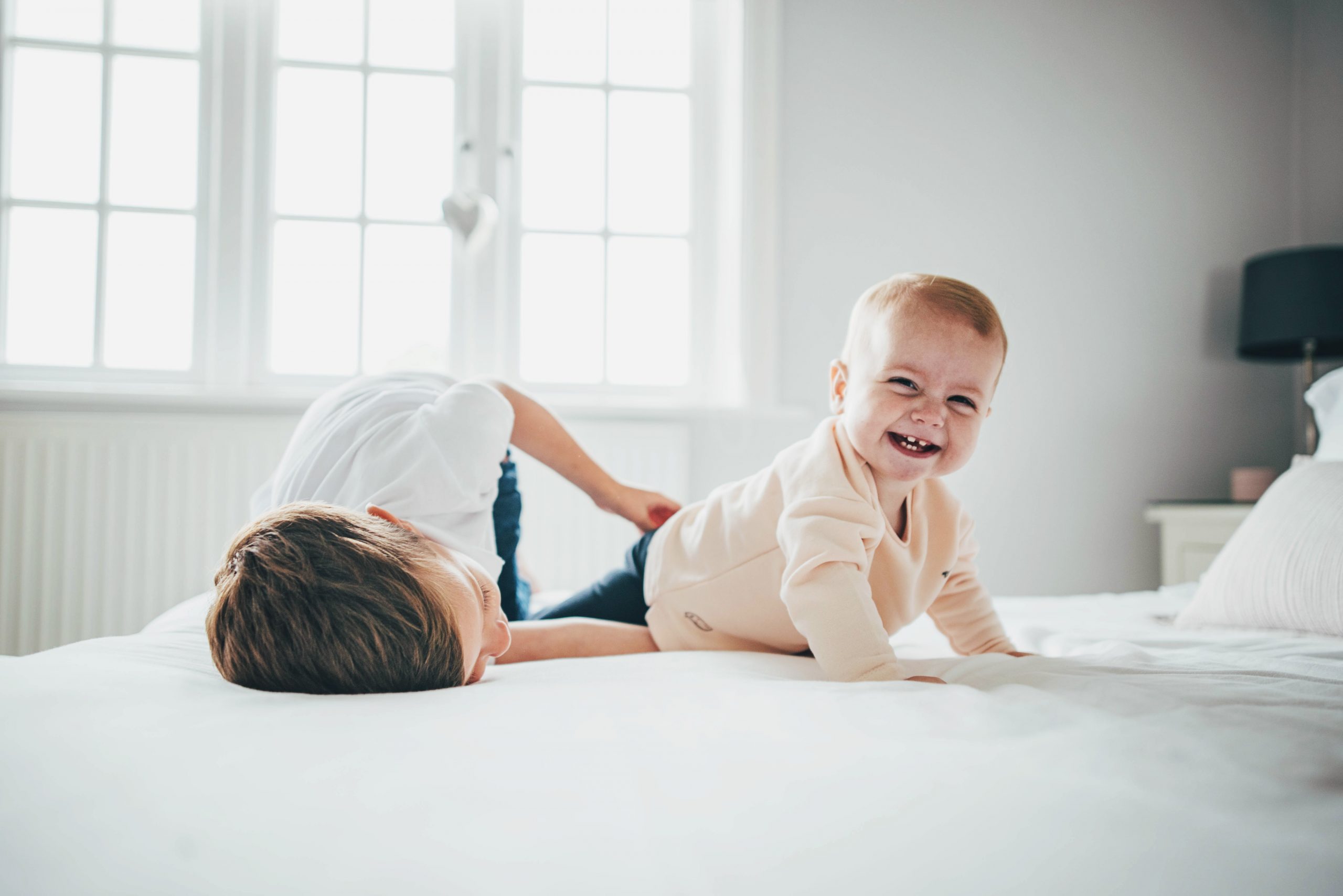 Little boy tickles baby sister on bed in white room