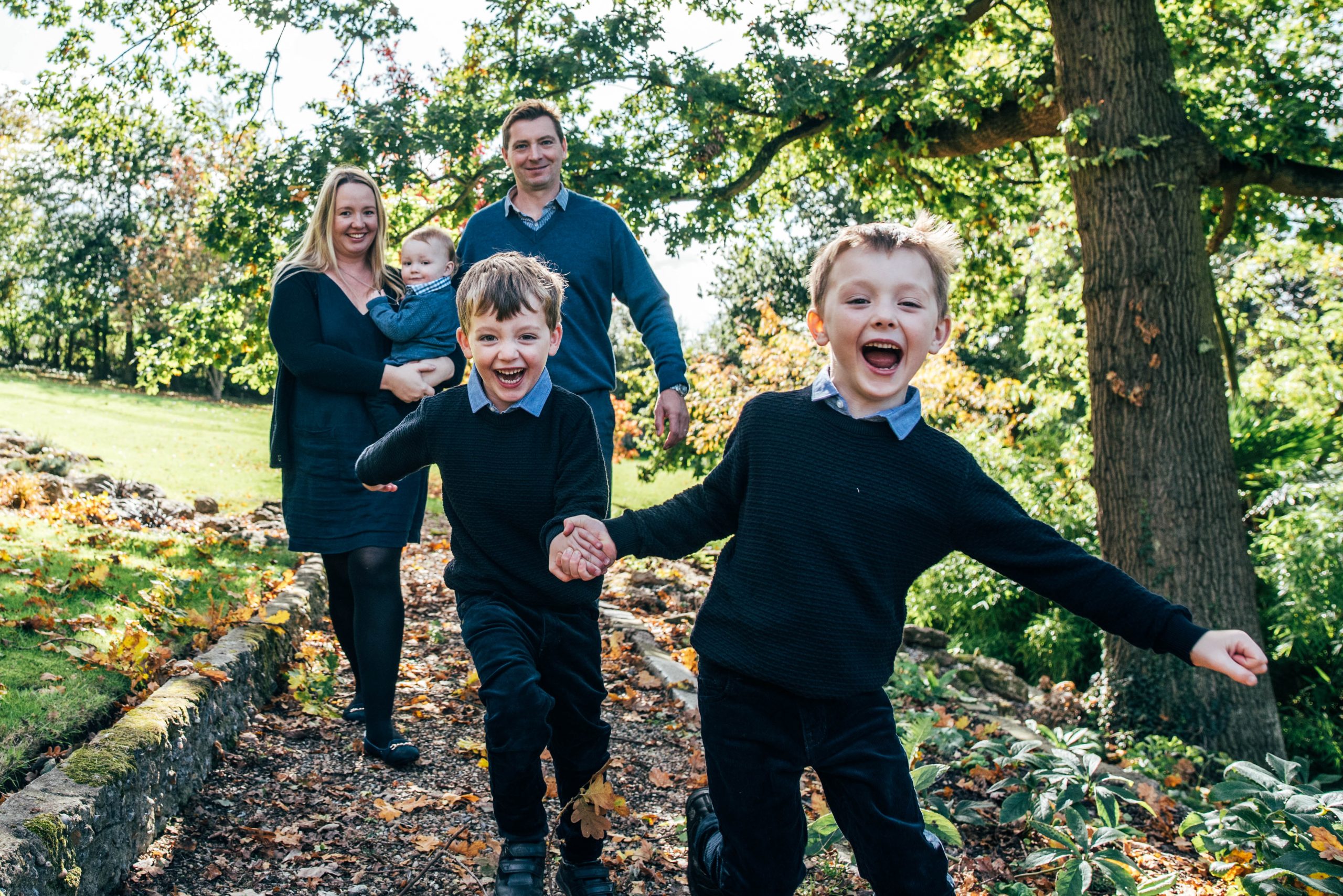 mum and dad hold baby as two older boys race towards the camera in garden in autumn