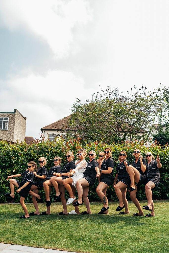 bride in white pyjamas and bridesmaids in black pyjamas do a TikTok routine in back garden whilst wearing heart sunglasses