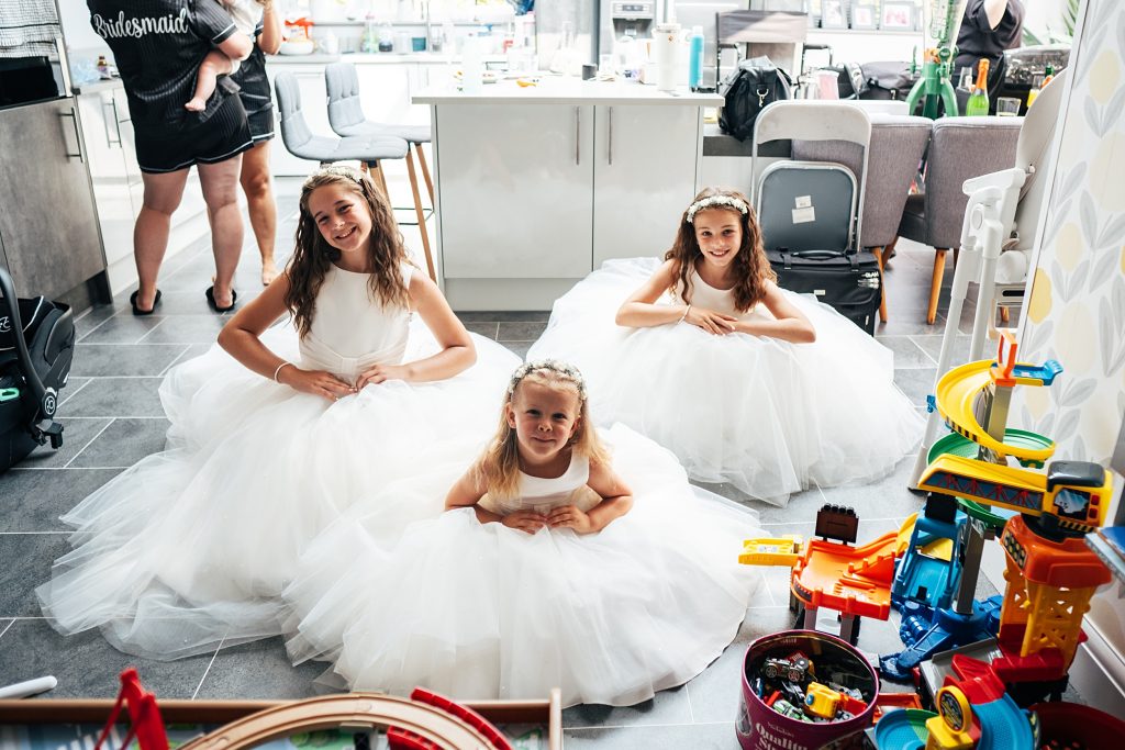 three flower girls in big tulle white dresses sit on floor surrounded by toys
