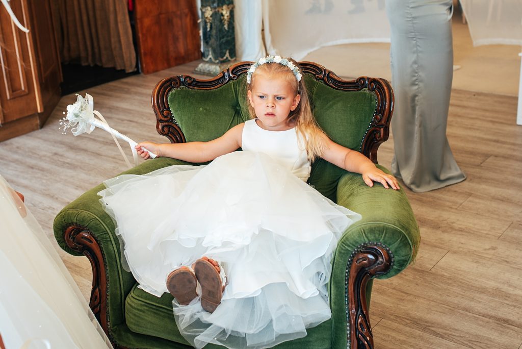 flower girl in white dress sits in a green velvet chair