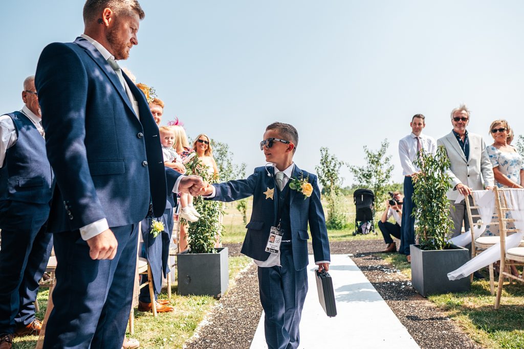 little boy in a suit and sunglasses carries a briefcase walks down aisle and fist bumps groom