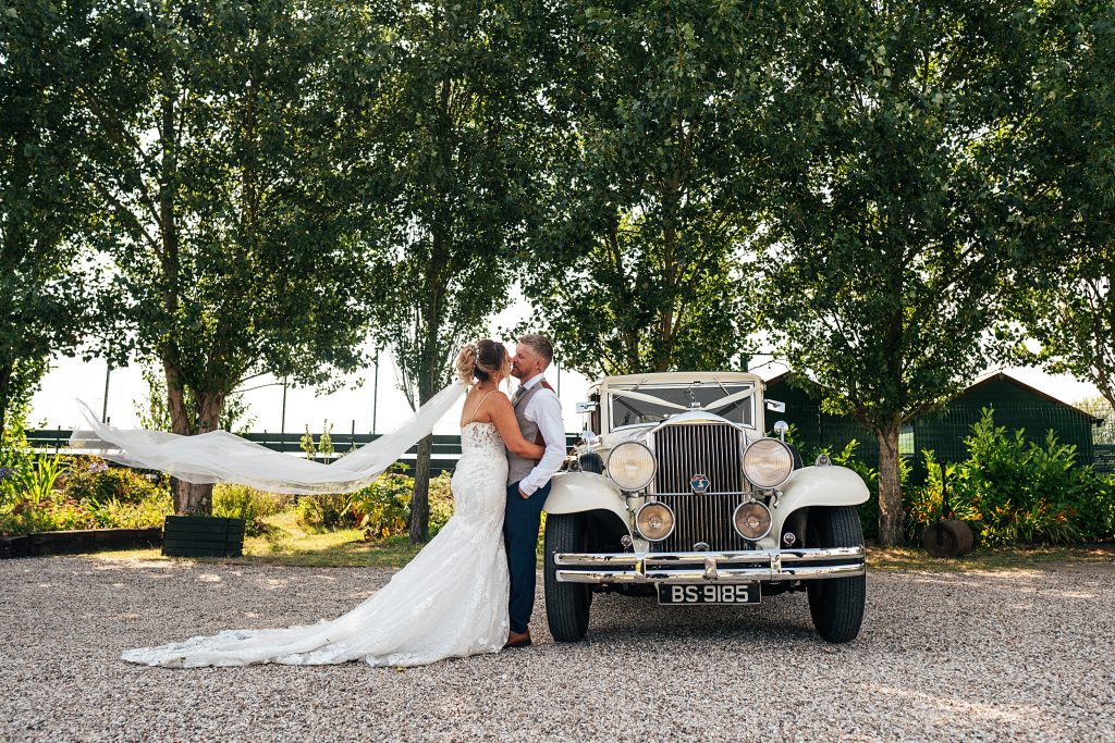 bride and groom kiss next to vintage car as veil billows in wind