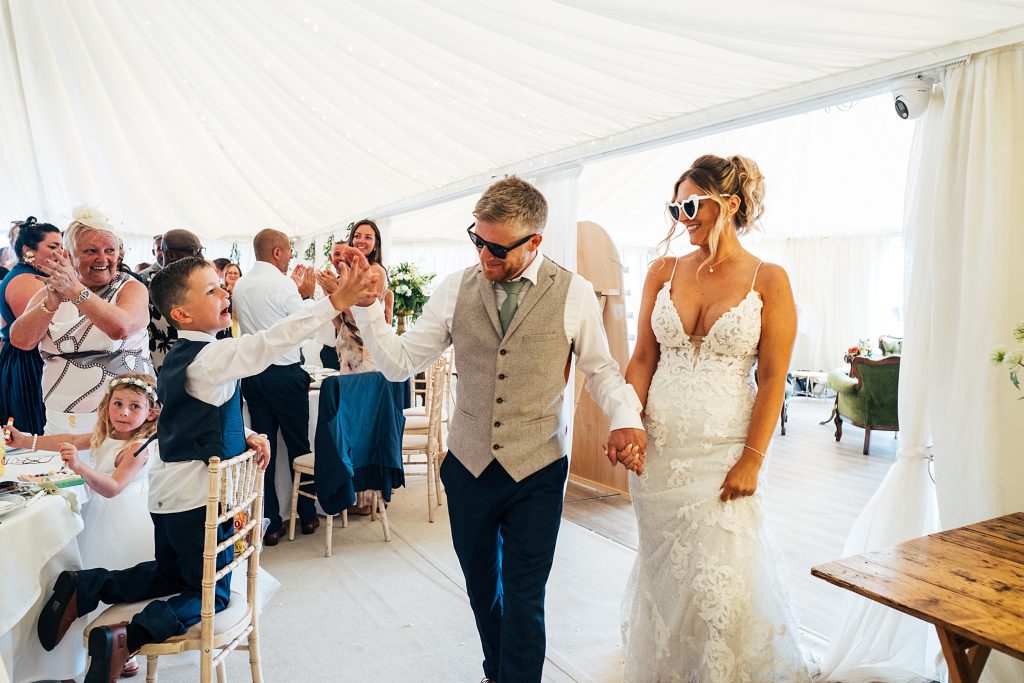 bride and groom enter marquee wearing sunglasses as groom fist bumps little boy