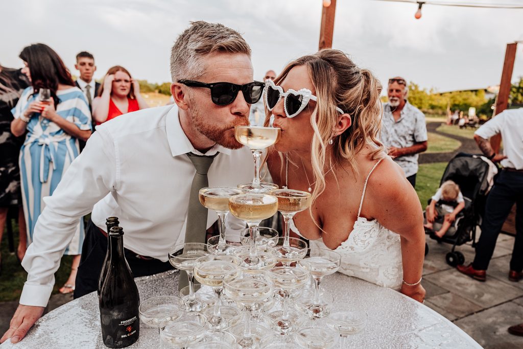 bride and groom in sunglasses sip from the top of a champagne tower