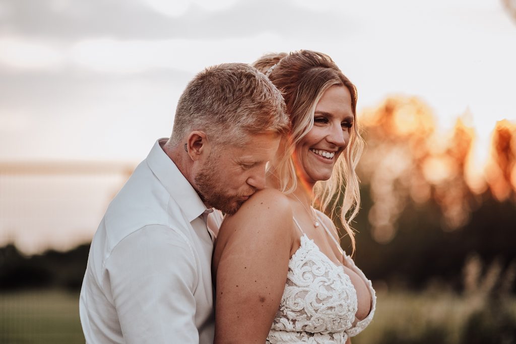 groom kisses brides shoulder at sunset